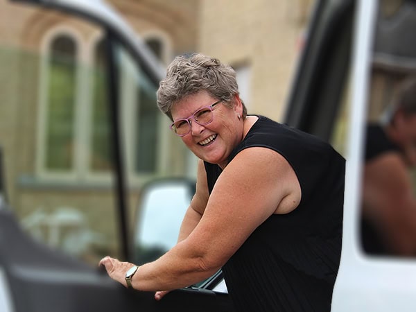 Een vrolijke persoon in zwarte kleding bij een auto, met een warme glimlach