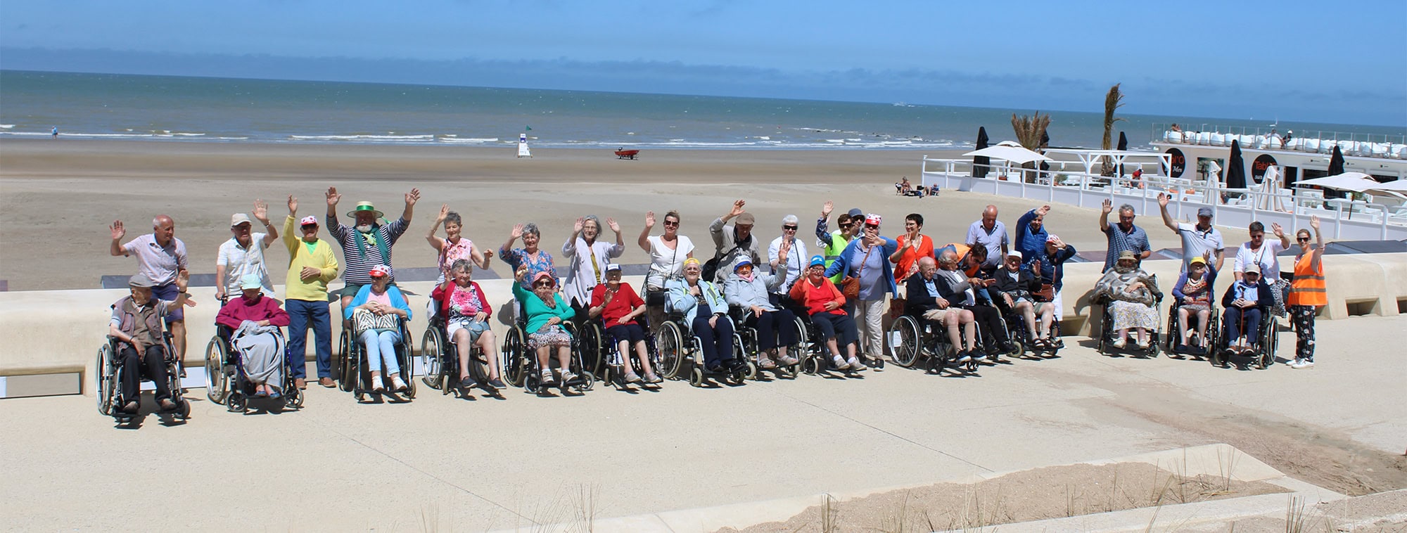 Groepsfoto van bewoners in rolstoelen met vrijwilligers op het strand, allemaal zwaaiend naar de camera
