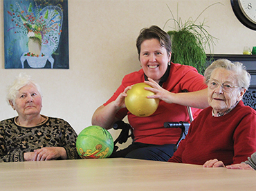 Beweegactiviteit met ballen in woonzorgcentrum, begeleider in rode trui tussen twee oudere dames
