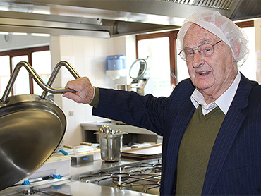 Oudere bewoner kijkt glimlachend in een grote kookpan in een professionele keuken