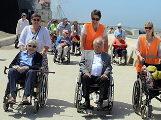 Vrijwilligers in oranje hesjes begeleiden bewoners in rolstoelen tijdens een wandeling op de boulevard