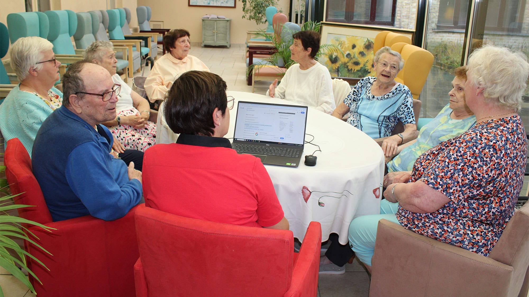 Een groep ouderen zit samen aan een ronde tafel en kijkt naar een laptop. Ze lijken deel te nemen aan een groepsactiviteit of bespreking in een gemeenschappelijke ruimte.
