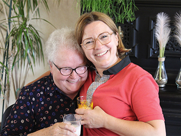 Twee vrouwen met brillen, een in rode trui en een in donkere blouse met print, glimlachend met drankjes in hun handen